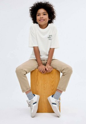 Bambino con capelli ricci seduto su una scatola di legno, indossa una maglietta bianca, pantaloni beige, calze grigie e scarpe da ginnastica bianche con strisce blu.