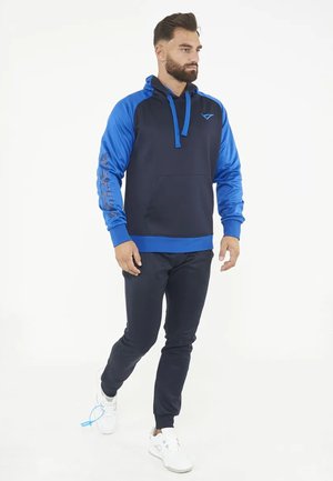Man loopt en draagt een marineblauw en blauw hoodie, zwarte broek en witte sneakers met blauwpaarse veters op één schoen.