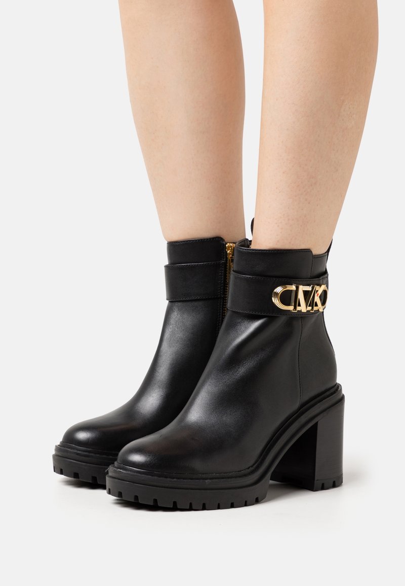 Bottines en cuir noir à talons hauts et épais, côtés avec fermeture éclair dorée et détails décoratifs de boucle dorée, portées sur des jambes nues.