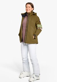 Olivgrüne Außenjacke mit Kapuze, ausgestattet mit farbigen Ärmelakzenten, getragen über einem lila Oberteil und weißen Schneehosen mit Skischuhen.