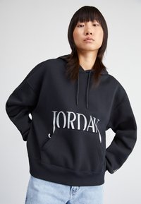 Sudadera con capucha negra y un bolsillo frontal. Presenta un gran texto "JORDAN" en plata. Corte holgado con puños y dobladillo acanalados. Textura de tela suave.