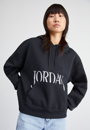 Sudadera con capucha negra y un bolsillo frontal. Presenta un gran texto "JORDAN" en plata. Corte holgado con puños y dobladillo acanalados. Textura de tela suave.