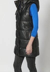 Femme portant une veste sans manches en cuir matelassé noire avec fermeture éclair et boutons-pression à l'avant, par-dessus un haut bleu à manches longues et un jean foncé.