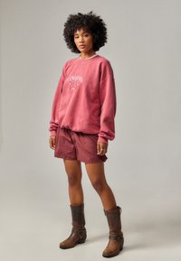 Rosa genser med grafisk design, kombinert med rød og hvit rutete shorts. Brune lærboots fullfører antrekket.