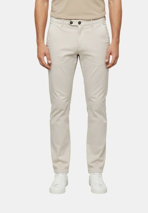 Pantaloni beige aderenti con due tasche anteriori, chiusura con doppio bottone nero, indossati con sneakers bianche e una camicia beige.