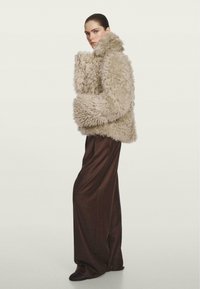 Puffige beigefarbene Kunstfelljacke mit hohem Kragen, kombiniert mit langen braunen Hosen. Die Jacke hat einen strukturierten, weichen Stoff und voluminöse Ärmel.