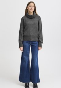 Maglione grigio scuro con collo a cowl in tessuto strutturato, abbinato a jeans a gamba larga blu. I jeans hanno un design classico a cinque tasche e un'abbellimento minimale.
