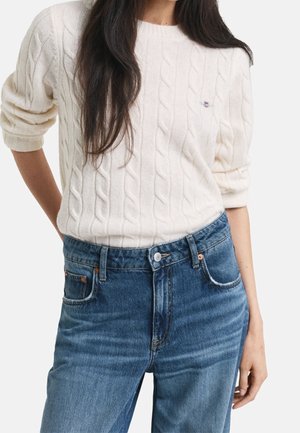 Femme portant un pull à torsades crème avec les manches retroussées et un jean bleu taille haute sur un fond uni.