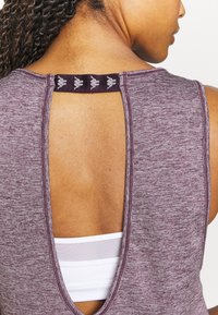 Haut sans manches couleur marron avec un dos ouvert. Présente un tissu texturé et une lanière noire avec un logo. Superposé à un soutien-gorge de sport blanc.