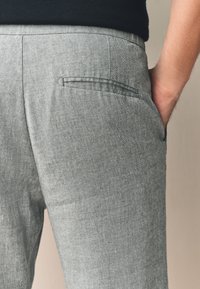 Šedé lněné kalhoty s elegantním střihem, hladkou texturou a jemným vzorem rybí kosti. Jedna zadní kapsa bez viditelných kovových prvků.