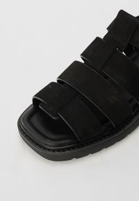 Sandalias de ante negras con múltiples correas anchas, puntera cuadrada y suela gruesa, que presentan costuras decorativas a lo largo de los bordes.