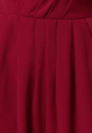 Vīna sarkanā auduma ar vertikālām locījuma krokām, kas veido plūstošu, teksturētu rakstu un maigus ēnas auduma virsmā.