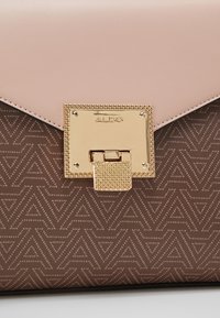 Zbliżenie brązowej torebki w wzory z różową klapką i złotym prostokątnym zapięciem z wygrawerowaną nazwą marki "ALDO".