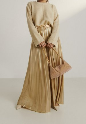 Femme portant un pull en tricot beige et une jupe plissée dorée brillante, tenant une pochette en maille dorée avec une sangle nouée.