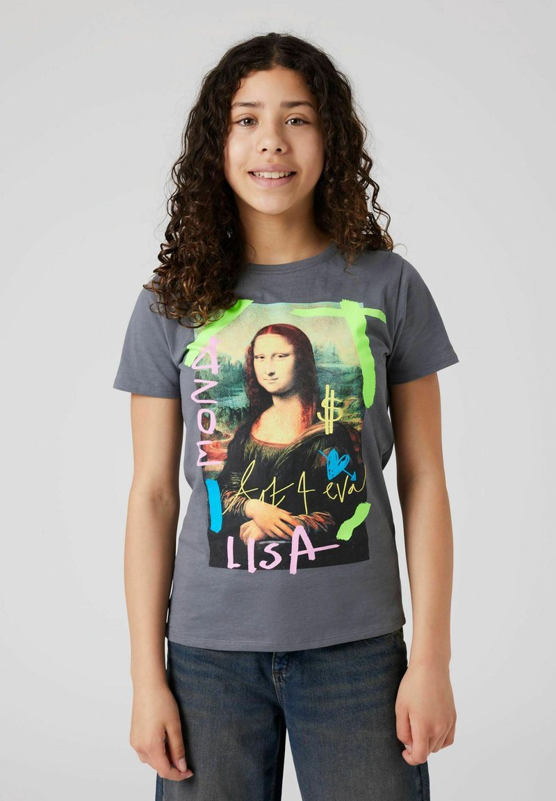 Meisje met krullend haar, gekleed in een grijze T-shirt met een kleurrijke graffiti-stijl Mona Lisa print en een blauwe spijkerbroek, staande tegen een effen achtergrond.