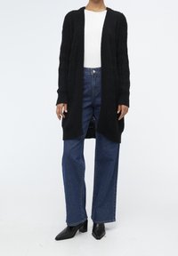Sort strikket cardigan med en løs pasform, der har et tekstureret mønster, lange ærmer og åben front. Sammen med højtaljede blå jeans og sorte hælede støvler.