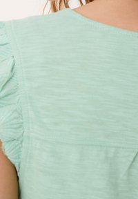 Světle zelená blůza s texturovanými volánovými rukávy. Vyrobena z měkké, prodyšné látky. Má kulatý výstřih a horizontální švy.