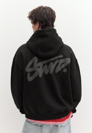 Jeune personne aux cheveux foncés portant un sweat à capuche noir avec un grand texte stylisé gris au dos, un jean clair et des chemises superposées en dessous.