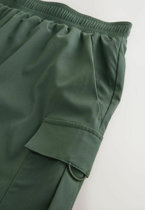 Oliivivihreät urheilushortsit, joissa on joustava vyötärö, sivutasku ja näkyvä rengas yksityiskohtana taskun läpässä. Sileä, kevyt kangas.