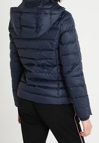 Tmavě modrá prošívaná péřová bunda s kapucí, s prošívanou texturou, elastickými manžetami a přiléhavým střihem. Vyrobena z lesklého, lehkého materiálu.