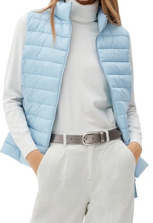 Světle modrá péřová vesta s vysokým límcem, vodorovným prošíváním a zipovým uzávěrem, nošená přes bílý rolák a světle šedé kalhoty.