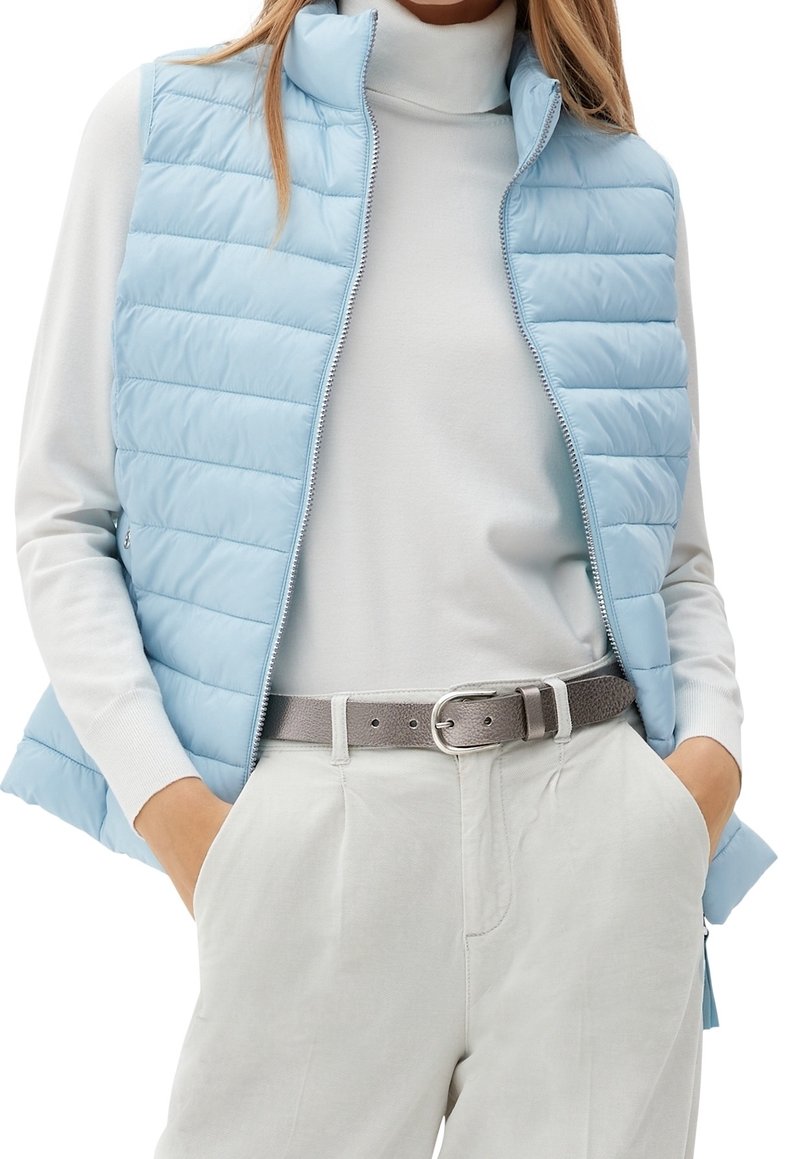 Světle modrá péřová vesta s vysokým límcem, vodorovným prošíváním a zipovým uzávěrem, nošená přes bílý rolák a světle šedé kalhoty.