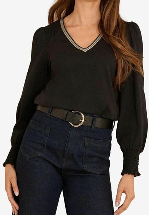 Nainen pukeutuneena mustaan V-kaulaiseen puseroon, jossa on liehuvat pitkät hihat, ja tummansinisiin farkkuihin, asusteena musta vyö, jossa on pyöreä kultainen solki.