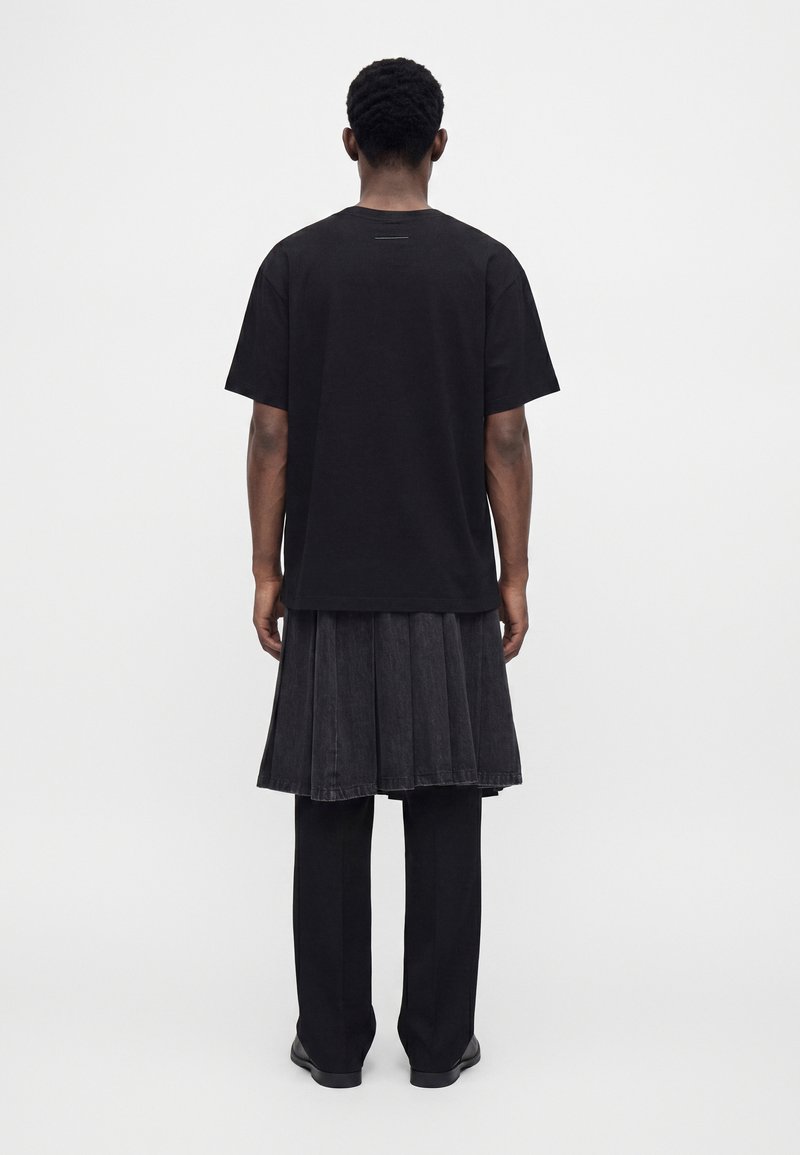 Person wearing a black short-sleeve shirt, black pleated skirt over black pants, standing facing away against a plain light background.