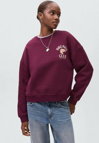 Giovane donna che indossa una felpa color bordeaux con la scritta "Brunch Club" e un grafico di un cornetto, abbinata a jeans blu chiaro e una collana d'argento.