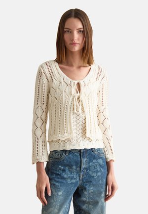 Woman wearing a cream crochet knit top with ties at the chest and floral patterned blue jeans, standing against a plain background.