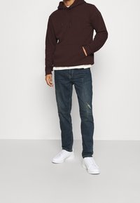 Sudadera marrón con un bolsillo canguro, combinada con jeans azules desgastados y zapatillas blancas. La sudadera tiene una capucha con cordón.