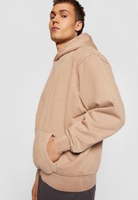 Bež hoodie iz bombaža s sprednjo kenguru žepom, rebrastimi manšetami in robom. Ima kapuco z vrvico in diskretno logo na prsih.