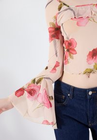 Haut à manches courtes en tissu beige transparent avec des manches évasées, orné de roses roses et de feuilles vertes. Associé à un jean bleu foncé.
