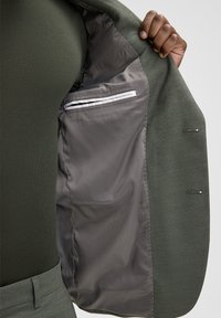 L'interno della giacca verde mostra una fodera grigia liscia con una tasca rifinita da un bordo bianco. Il tessuto esterno presenta una finitura testurizzata.