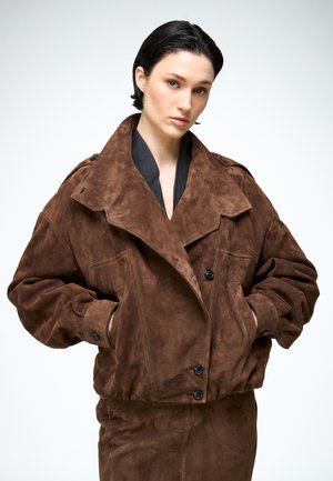 Junge Frau mit kurzen dunklen Haaren, die eine braune, übergroße Wildlederjacke und einen passenden Rock trägt, mit den Händen in den Taschen stehend.