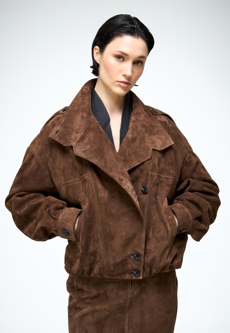 Jonge vrouw met kort donker haar, gekleed in een oversized jasje van bruin suède en een bijpassende rok, staand met haar handen in de zakken.