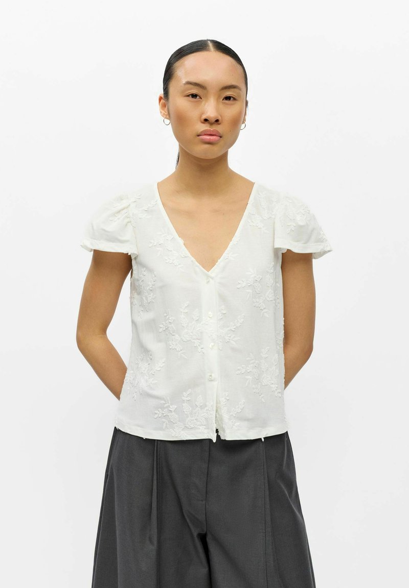 Jeune femme aux cheveux foncés attachés porte un chemisier blanc texturé à manches courtes avec un motif floral et un pantalon ample gris foncé, debout sur fond blanc.