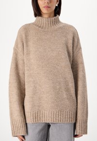 Beige turtleneck sweater with ribbed hem and cuffs. Soft texture, relaxed fit, and dropped shoulders. Worn over gray pants.