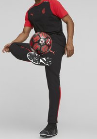 Conjunto deportivo de Puma con una camiseta de manga corta negra y roja, pantalones negros con acentos rojos y botas de fútbol negras, equilibrando un balón de fútbol con un patrón.