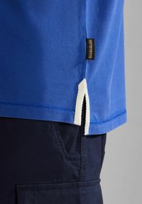 Tessuto di una polo blu con un motivo testurizzato, dotata di uno spacco laterale con finiture bianco sporco e una piccola etichetta del marchio sull'orlo.