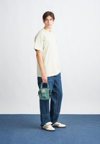 Svetlošedé oversized tričko, modré voľné jeansy, biele tenisky s čiernymi pruhmi, drží malú zelenú kabelku s logom.