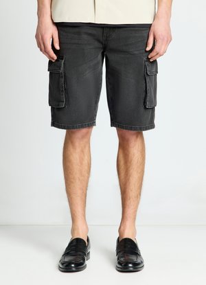 Uomo in piedi che indossa pantaloncini cargo in denim nero con tasche laterali e scarpe slip-on in pelle nera su uno sfondo chiaro e semplice.