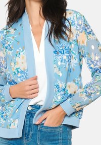 Květinová bomber bunda ve světle modré barvě, se bílými a zlatými akcenty, s žebrovanými manžetami a spodním lemem. Nosená přes bílý top a džíny.