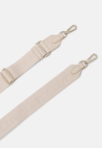 Beige textilní pásek Calvin Klein s embosovaným logem, vybavený kovovými sponami a akcenty ve zlatém tónu, plochý design a minimalistický styl.