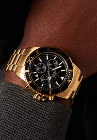 Goldene Edelstahl-Armbanduhr mit schwarzem Ziffernblatt, weißen Markierungen, mehreren Unterzifferblättern und strukturiertem Armband, das an einem Handgelenk positioniert ist.