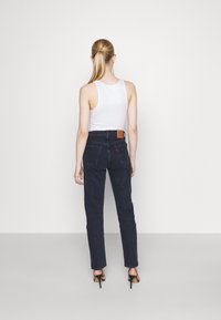 Jeans de mezclilla de color azul oscuro con un diseño ajustado, cintura alta y un parche de cuero marrón en la parte trasera, combinados con una camiseta sin mangas blanca y tacones.