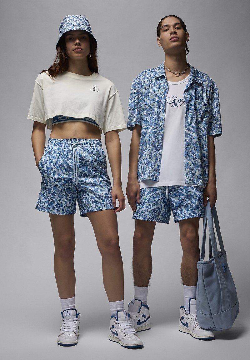 Deux modèles portent des tenues assorties à motif camouflage bleu : un top beige court, un short bleu et un chapeau seau ; le modèle masculin porte également une chemise à boutons et un tote.