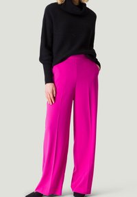 Suéter negro de punto con cuello alto, combinado con pantalones de pierna ancha en un brillante color rosa. Los pantalones tienen una textura suave y un corte relajado.