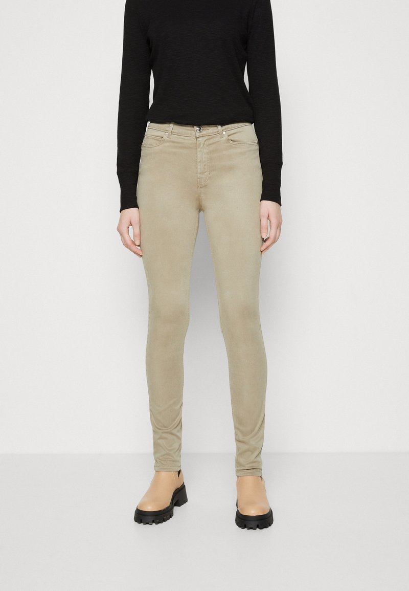 Personne portant un pantalon slim beige, un haut à manches longues noir et des bottes beige à semelle épaisse, debout devant un fond blanc uni.