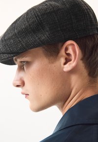 Siva kapa s uzorkom pasjeg zuba, izrađena od teksturirane tkanine, s zakrivljenim vrhom. Model ima kratku kosu, vidljivo uho i čistu liniju čeljusti.